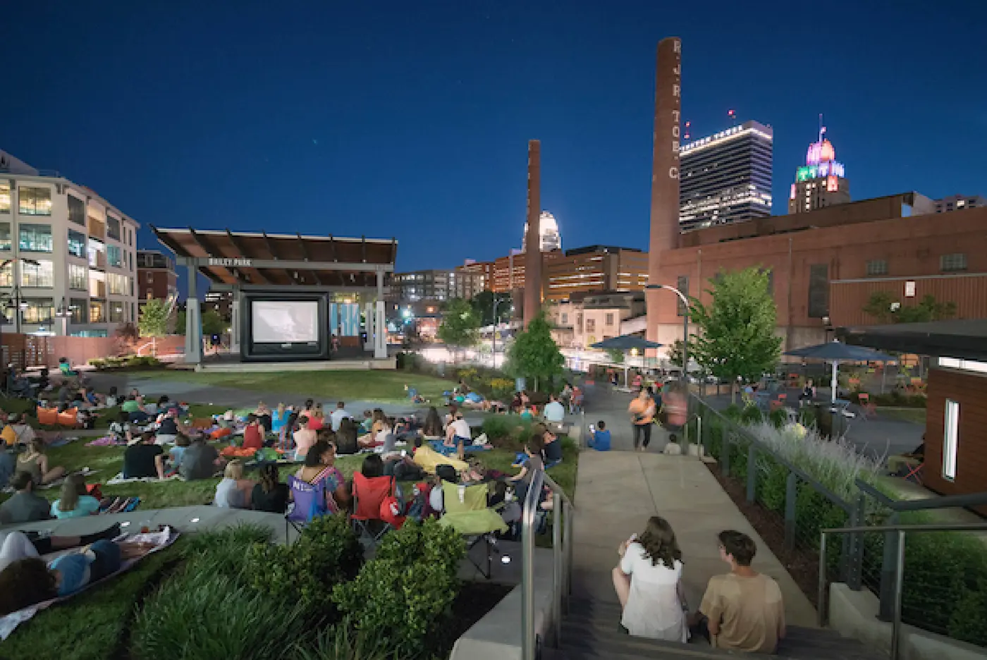 A crowd gathered on the lawn at Bailey Park for an outdoor movie at dusk, with the Winston-Salem skyline in the background.